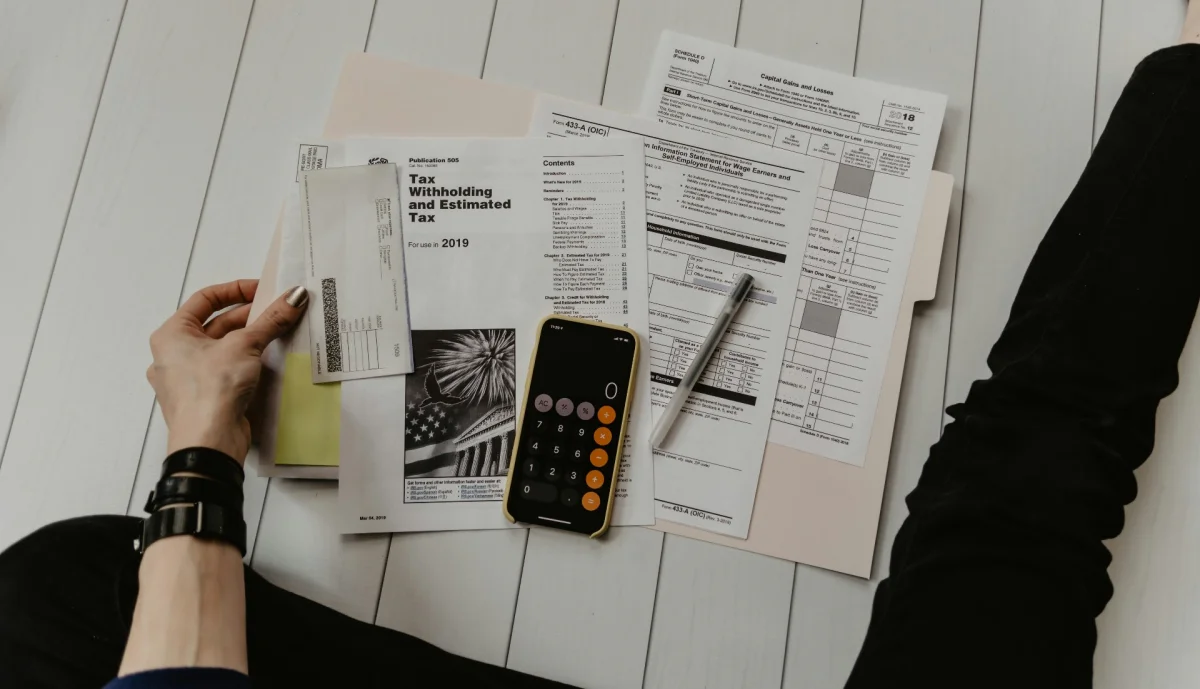 Tax forms, a phone calculator and a pen spread on a desk, with a hand reaching for a document