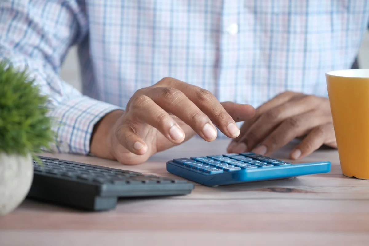 Hands using a calculator on a desk beside a keyboard and a mug