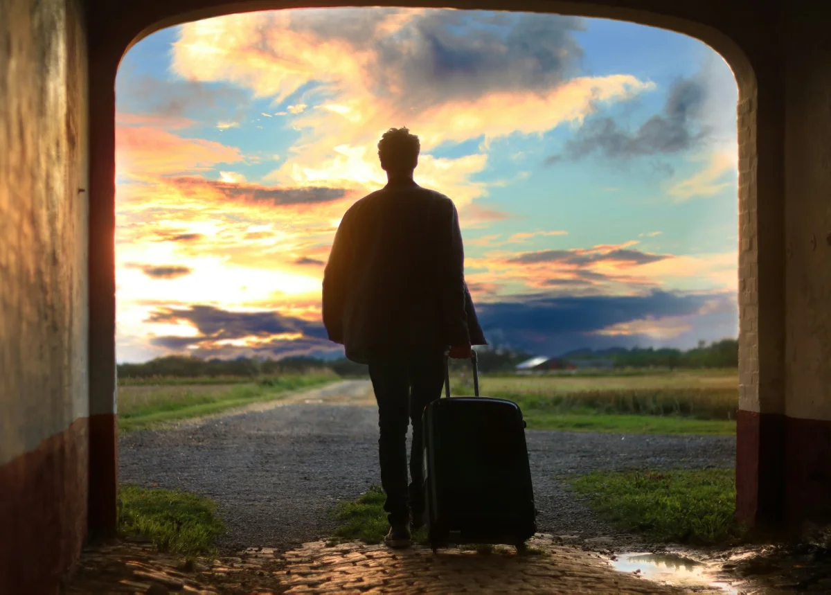 Person with a rolling suitcase standing in a doorway, facing a road and a sunset sky