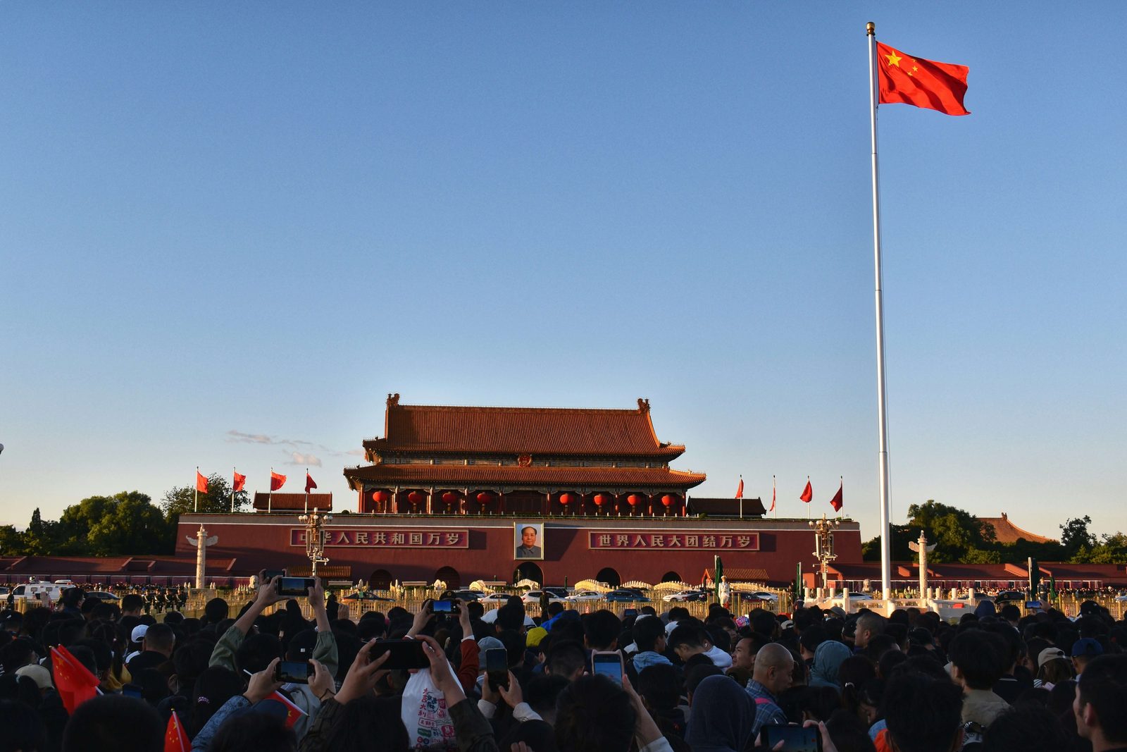 A crowd at Tian'anmen at dawn holding up phones and small flags toward the national flag on its pole
