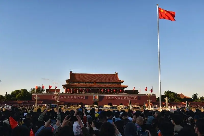A crowd at Tian'anmen at dawn holding up phones and small flags toward the national flag on its pole