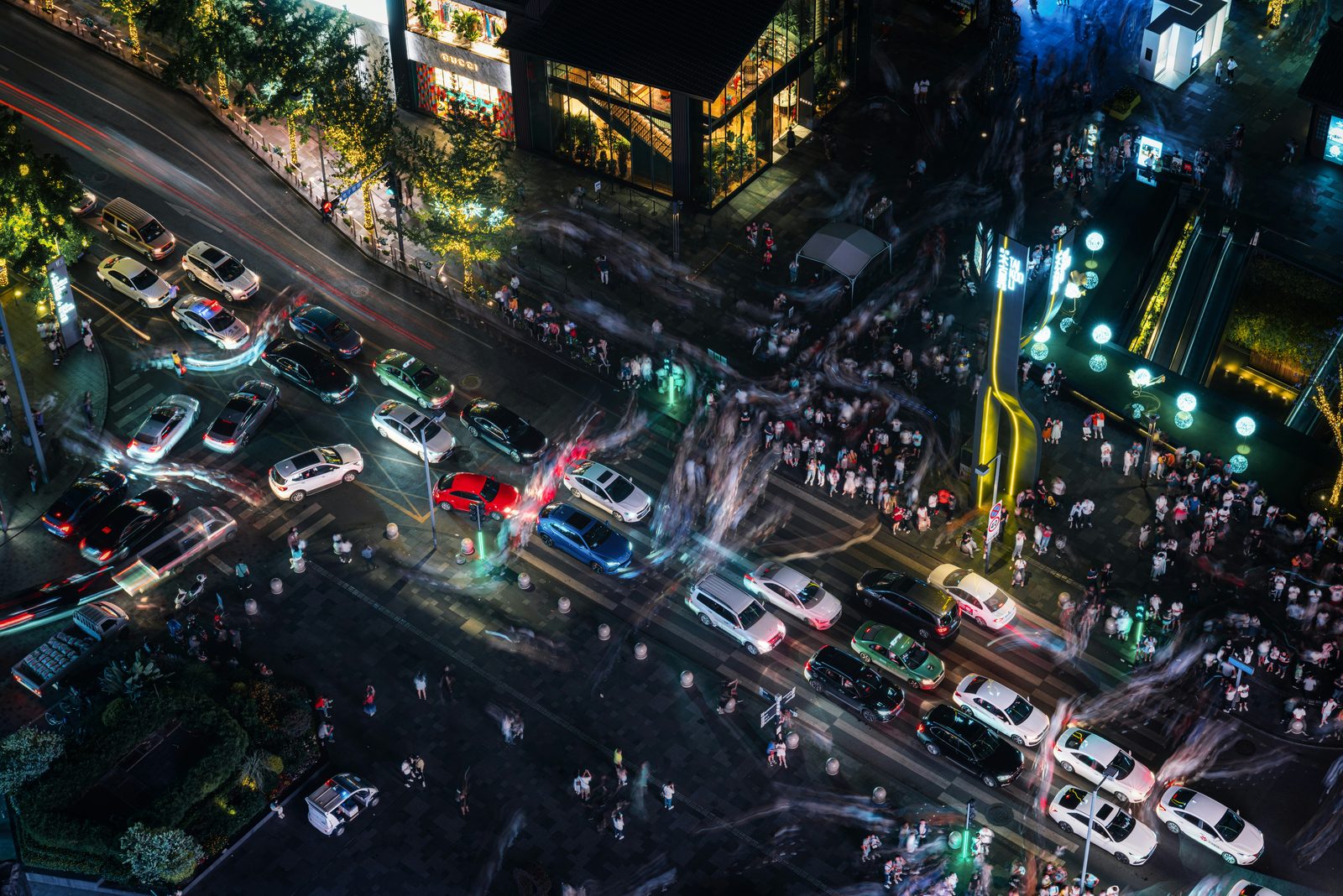 A Chengdu intersection at night seen from above — dense traffic, luxury storefronts and blurred crowds streaming across the crosswalks
