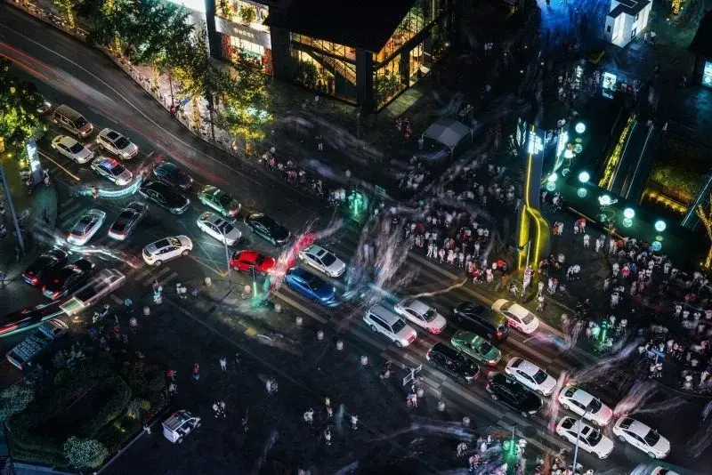 A Chengdu intersection at night seen from above — dense traffic, luxury storefronts and blurred crowds streaming across the crosswalks
