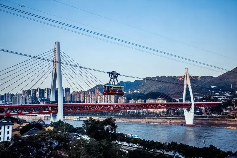 A red cable car of the Yangtze Ropeway crossing the river in Chongqing, suspended between two white pylons with the city skyline behind