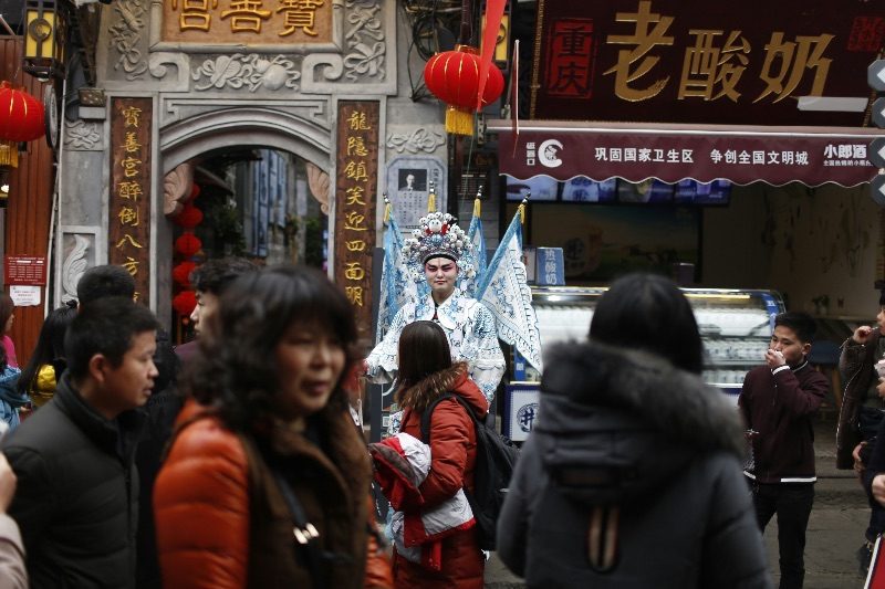 A costumed performer in traditional opera dress parades through a crowded lane in Ciqikou old town, Chongqing, past lantern-strung shopfronts