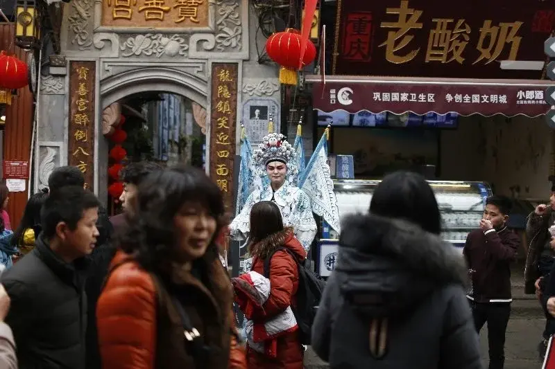 A costumed performer in traditional opera dress parades through a crowded lane in Ciqikou old town, Chongqing, past lantern-strung shopfronts