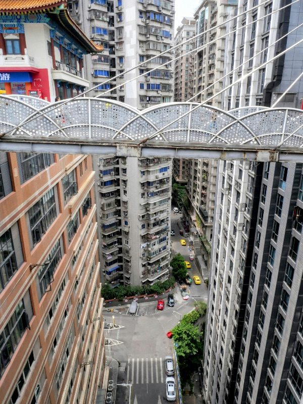 Looking down from the Kuixing Lou skybridge in Chongqing at a residential tower built down a hillside, with a red-and-gold pavilion rooftop visible at the same level as the bridge deck