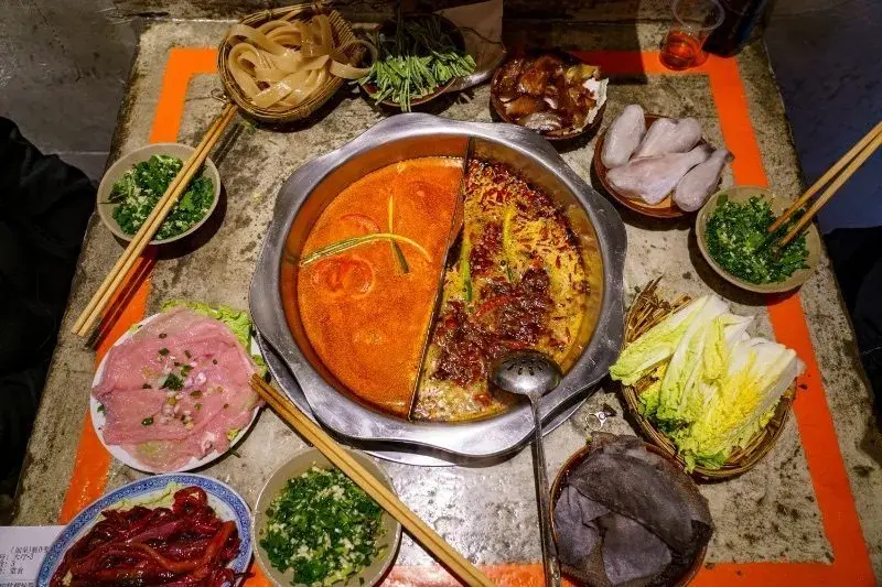 An overhead view of a divided Chongqing hotpot, orange broth on one side and dark chili broth on the other, surrounded by small dishes of raw ingredients