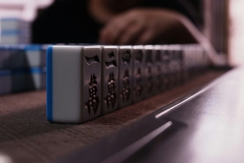 A close-up row of mahjong tiles lined up on a table, a player's hand blurred in the background