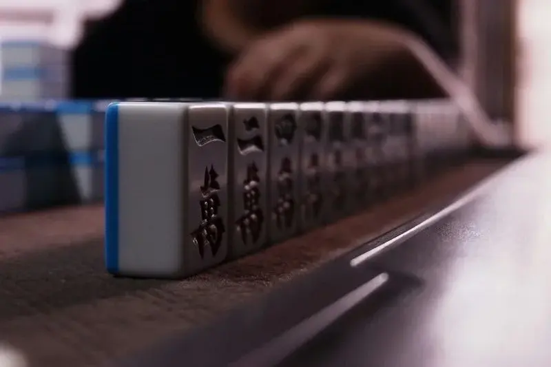 A close-up row of mahjong tiles lined up on a table, a player's hand blurred in the background