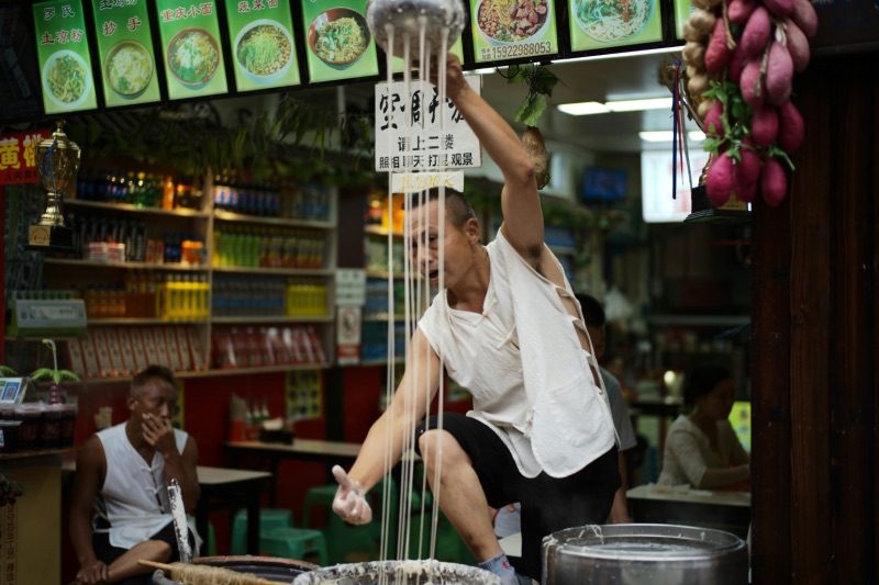 A street vendor hand-pulling noodles at a Chongqing noodle stall, dough stretched overhead