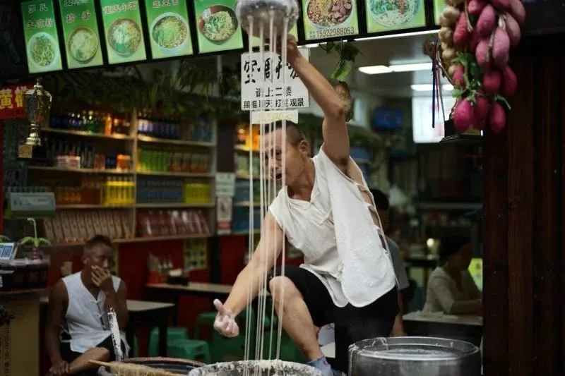 A street vendor hand-pulling noodles at a Chongqing noodle stall, dough stretched overhead