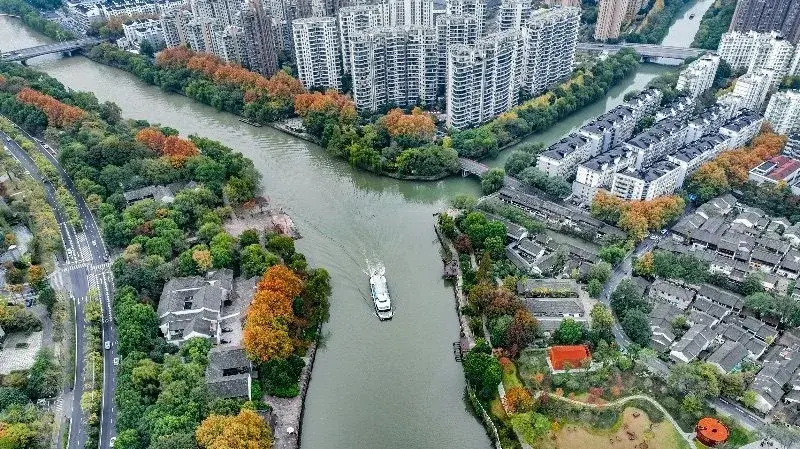 Aerial view of a Hangzhou canal, a boat passing between a dense high-rise apartment complex on one bank and low traditional tile-roofed houses on the other, autumn trees along the water