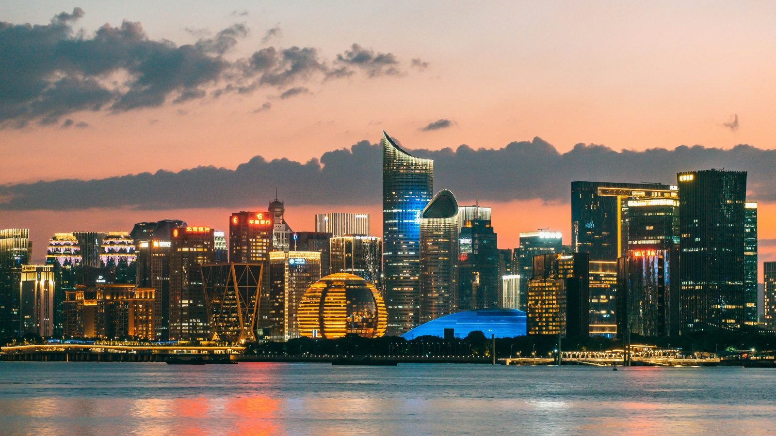 Hangzhou's Qianjiang New Town skyline at sunset, the golden ring-shaped tower lit up beside the river, dramatic clouds overhead