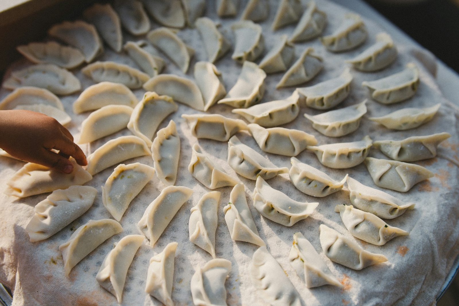 Dozens of raw dumplings arranged on a cloth-lined tray, a child's hand reaching toward one