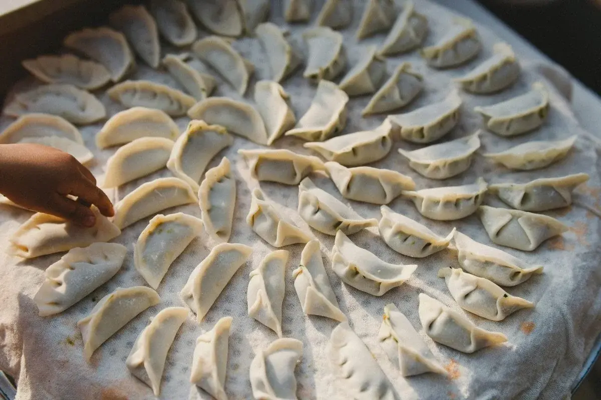 Dozens of raw dumplings arranged on a cloth-lined tray, a child's hand reaching toward one