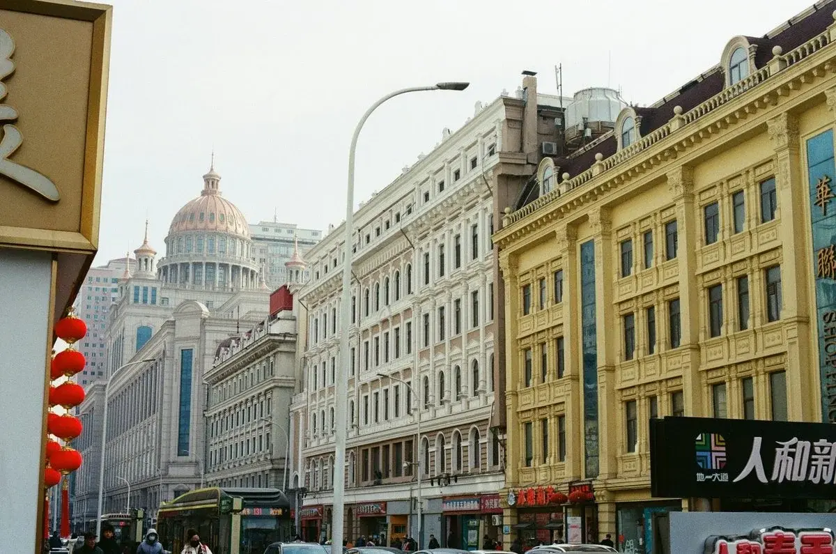 A Harbin street lined with ornate European-style buildings and a copper dome, red Chinese lanterns and shopping signage in the foreground