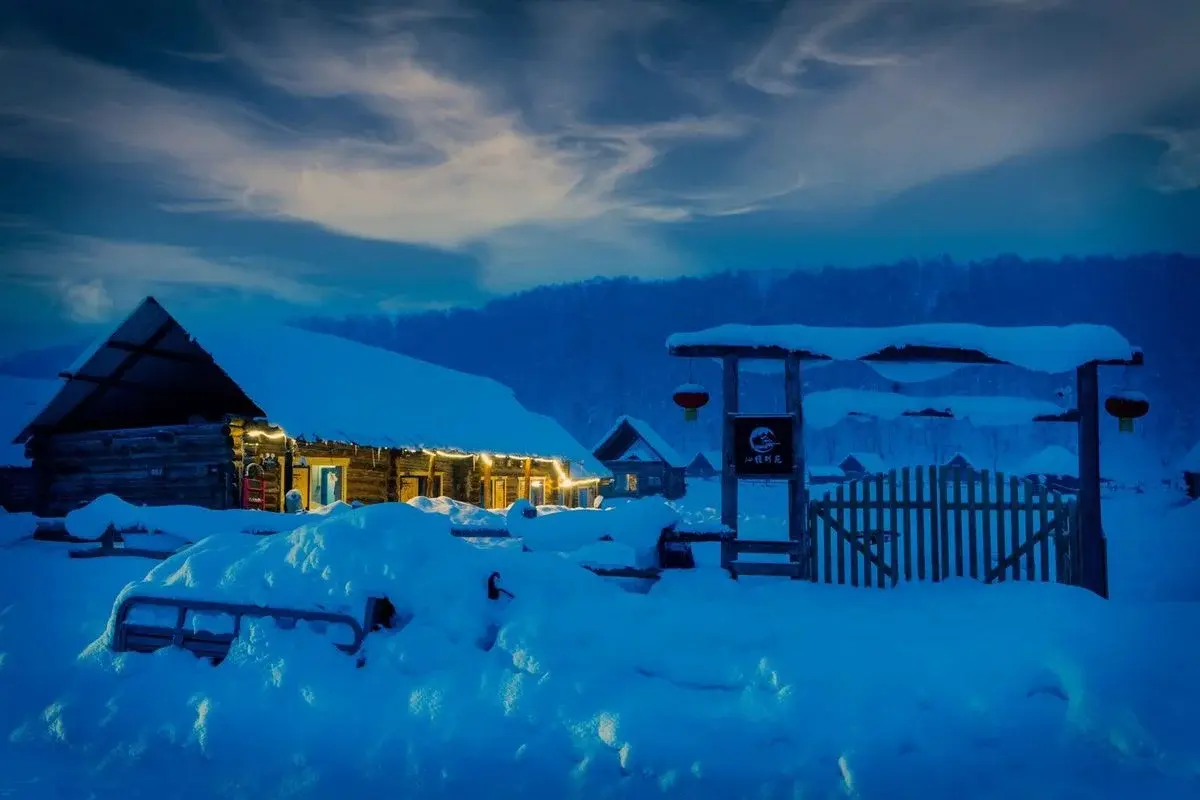 A snow-buried village at blue hour, log cabins under a metre of snow with warm light in the windows