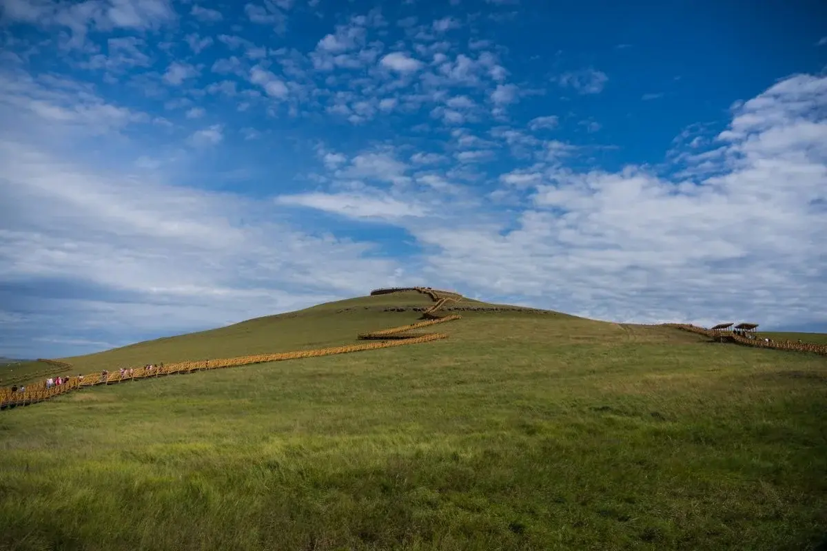 A grassy hill with a wooden boardwalk winding to the summit under a wide blue sky with scattered white clouds near Hohhot
