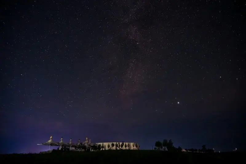 A dark starry sky with the Milky Way visible over a lit yurt camp on the grassland at night