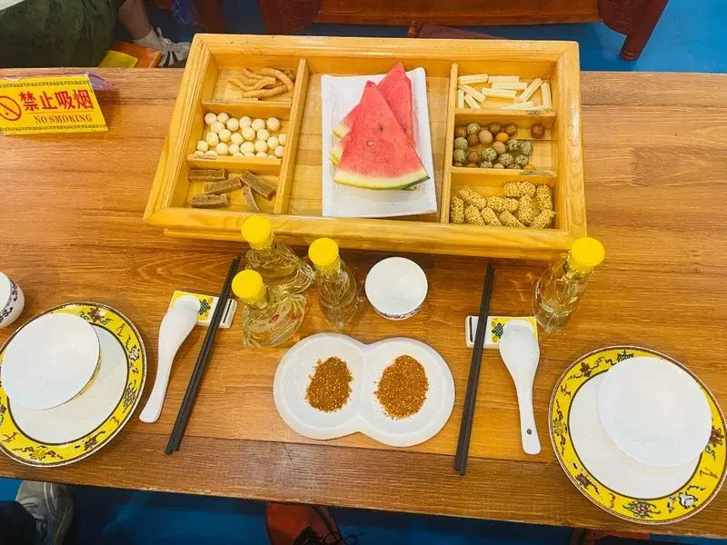 A feast-hall table set with bottles of baijiu, dried snacks, watermelon, and dipping condiments for a zhamayan banquet