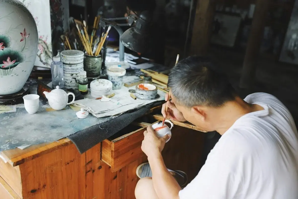 A craftsman's hands carving flower petals into the surface of a raw clay pot with a metal tool
