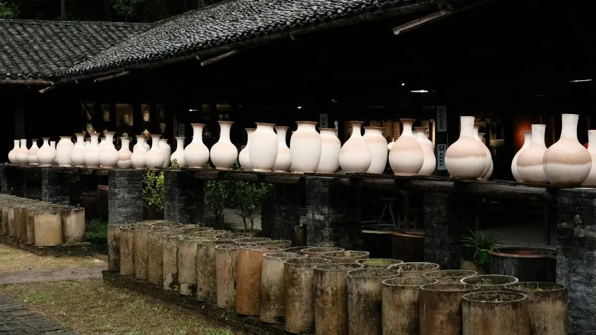 Rows of unfired white porcelain vases drying on a rack above rows of old ceramic saggars at a historic kiln site