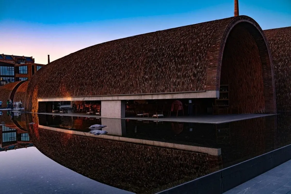 The curved brick shell of Taoxichuan at dusk, a modern building made from an old porcelain factory, reflected in still water