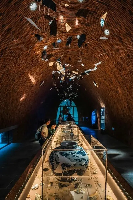 A museum gallery built inside a curved brick kiln-shaped hall, a blue-and-white porcelain jar in a glass case beneath a ceiling installation of suspended porcelain shards