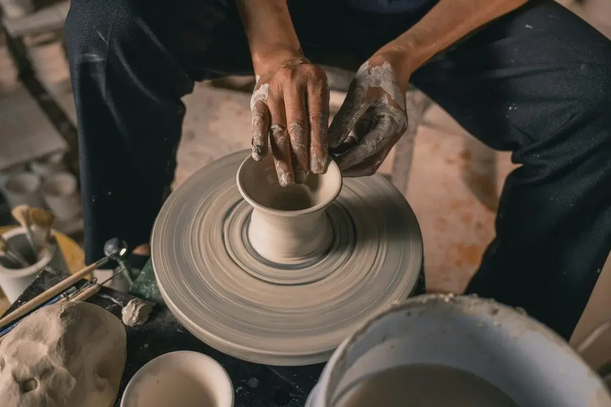 A potter's mud-caked hands shaping the walls of a spinning cup on a wheel, seen from above