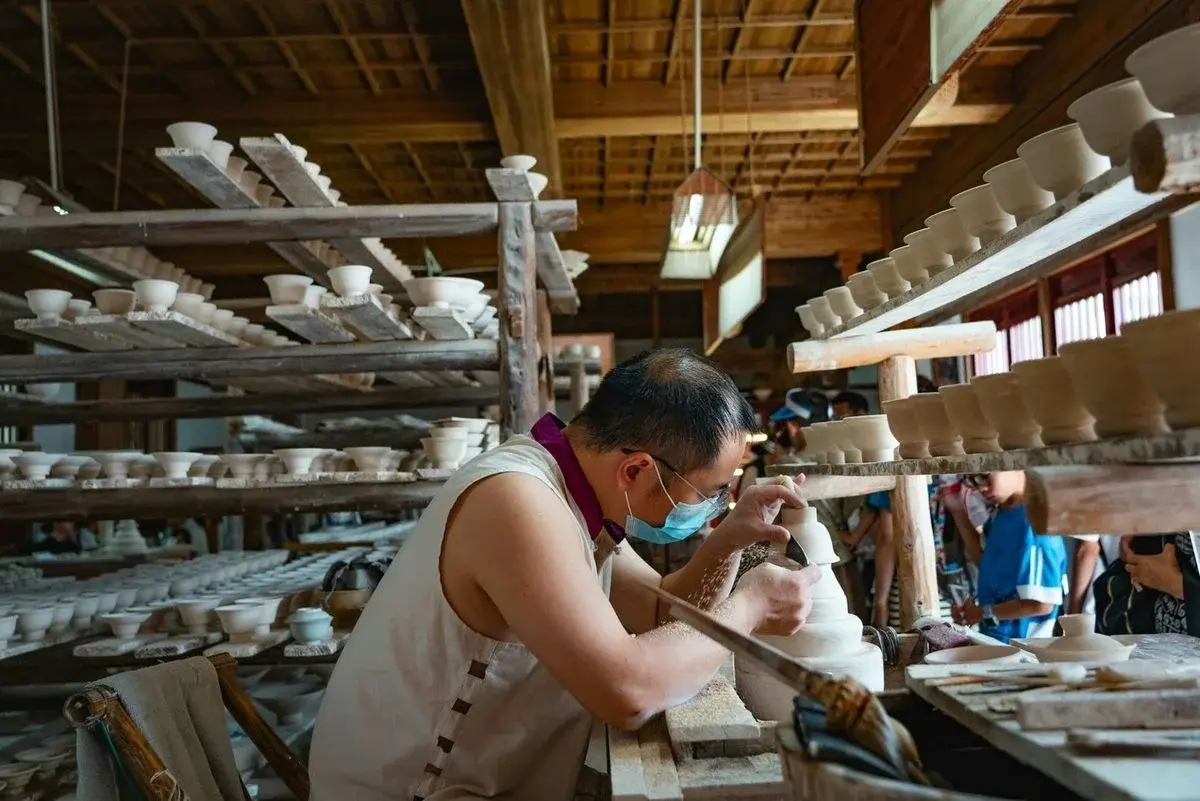 A masked craftsman trimming a cup at a workbench, surrounded by wooden racks holding hundreds of pale unfired pieces, tourists watching in the background