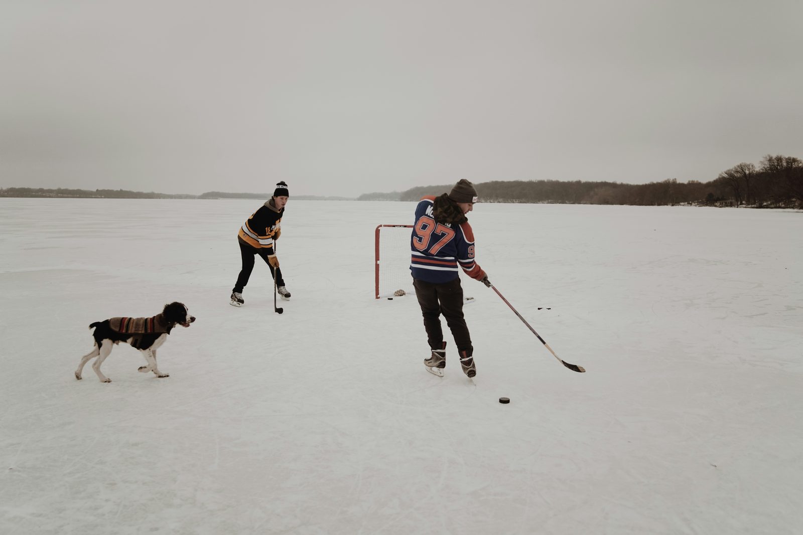 Two people playing hockey on a frozen lake with a dog watching