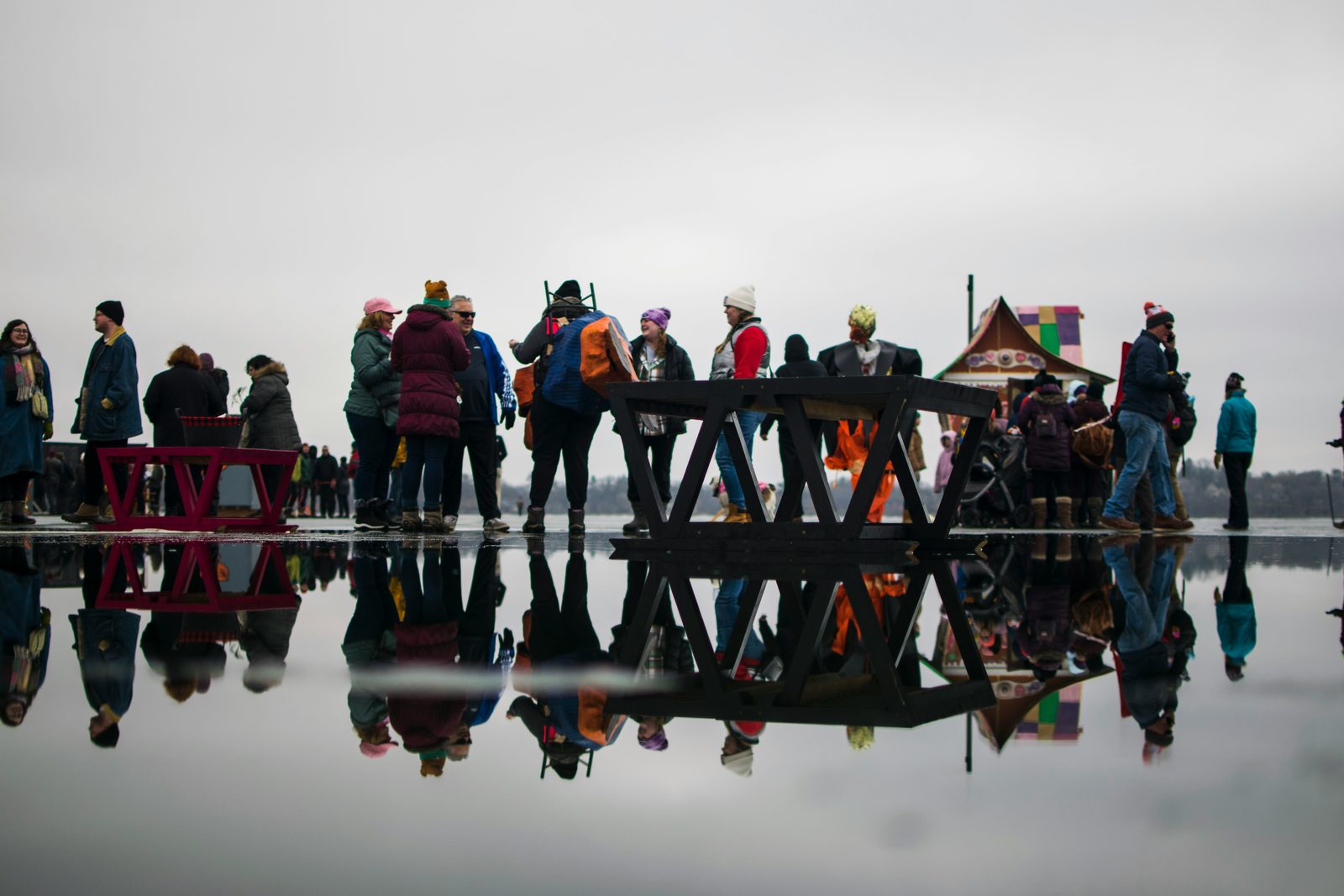 People gathered on a frozen Minnesota lake around wooden structures, reflected in the ice