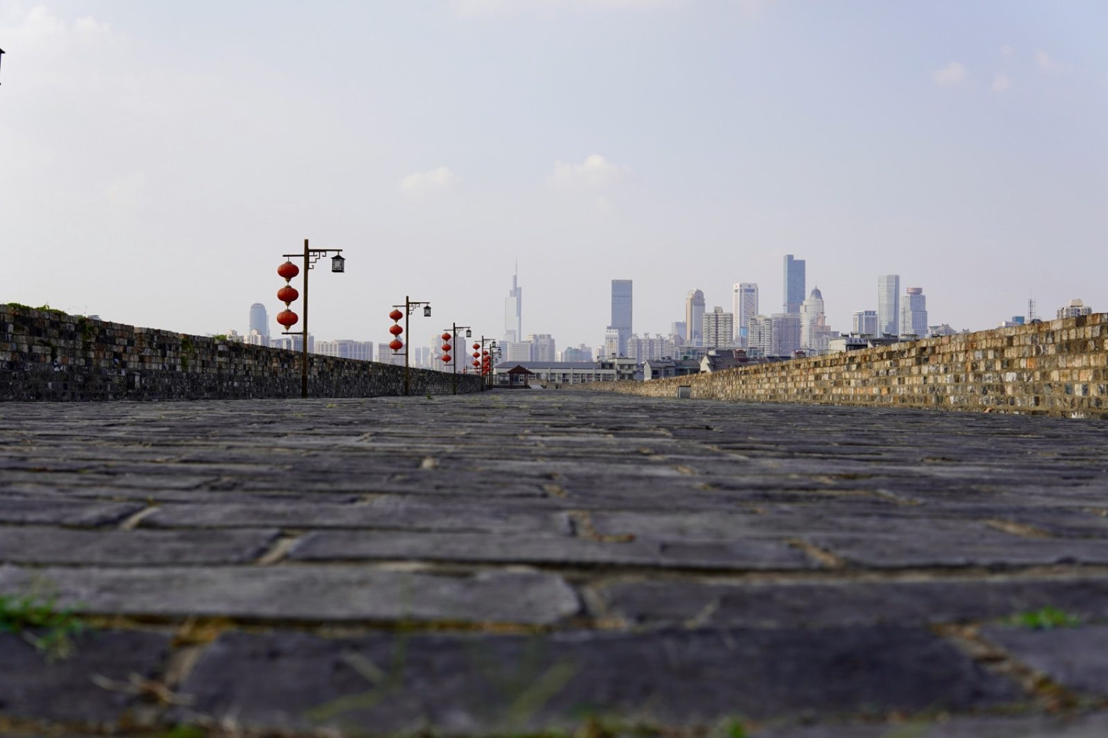 Weathered brick atop the Ming-dynasty city wall of Nanjing, red lanterns lining the walkway, with the modern skyline visible in the distance