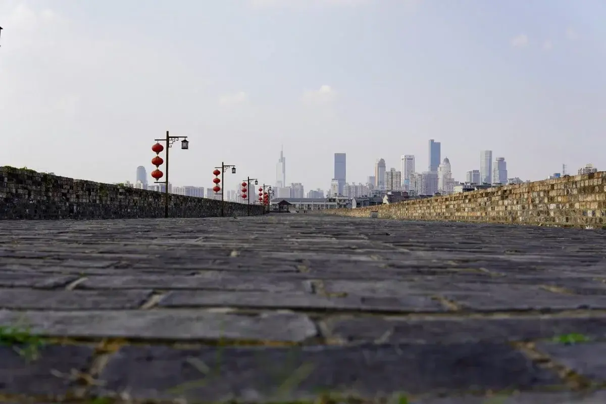 Weathered brick atop the Ming-dynasty city wall of Nanjing, red lanterns lining the walkway, with the modern skyline visible in the distance