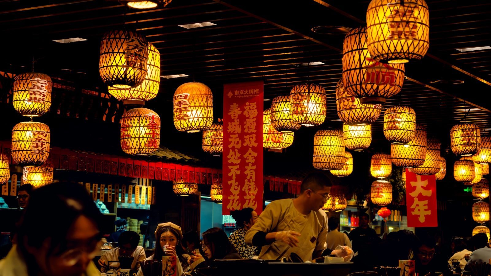 Rows of glowing lanterns hanging inside a busy Nanjing Impressions restaurant at night, with red banners and diners seated below