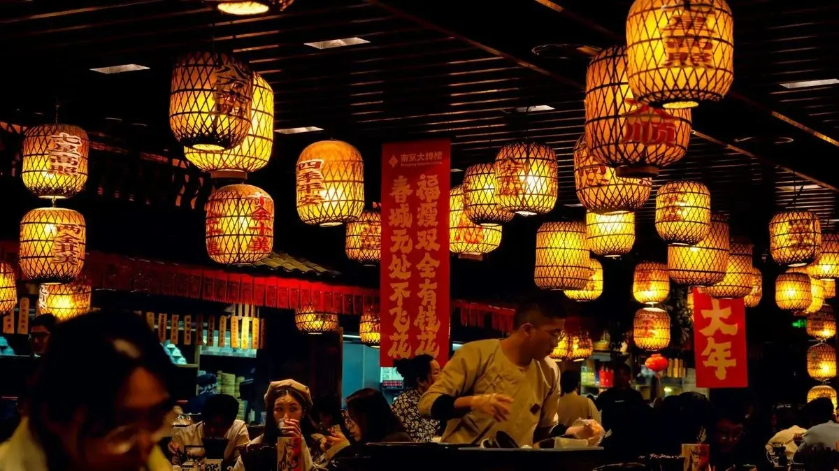 Rows of glowing lanterns hanging inside a busy Nanjing Impressions restaurant at night, with red banners and diners seated below