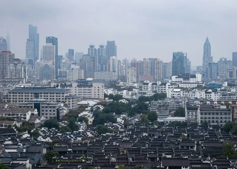 A dense grid of low tiled-roof traditional houses in the foreground, with modern high-rise towers and a hazy skyline rising behind them in Nanjing