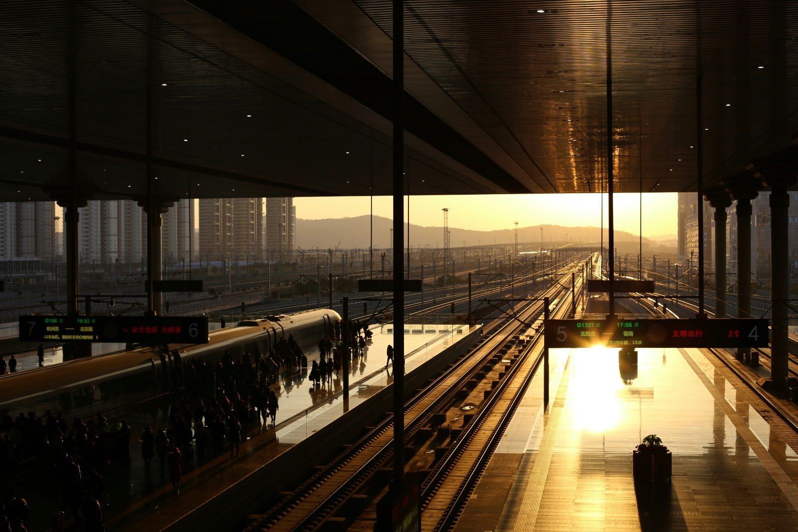 A high-speed rail platform at sunset, a train pulling in as passengers wait, departure boards showing routes to Hangzhou East, Beijing South and Shenyang North