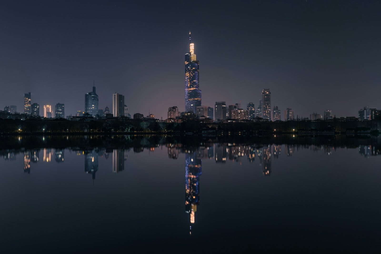 The Zifeng Tower lit up at night, reflected in a still lake alongside the rest of Nanjing's financial-district skyline