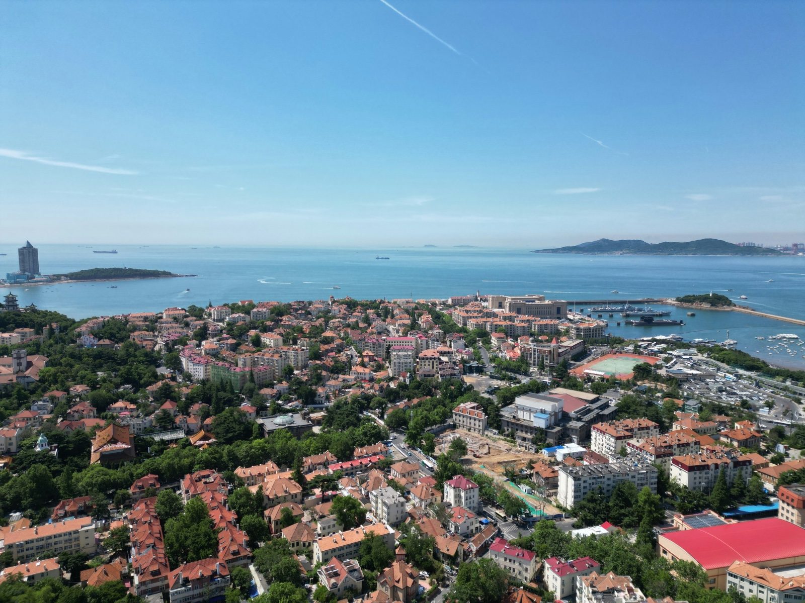 Aerial view of Qingdao's red-tile-roofed old town spread along a curving harbor, boats moored offshore and green hills behind