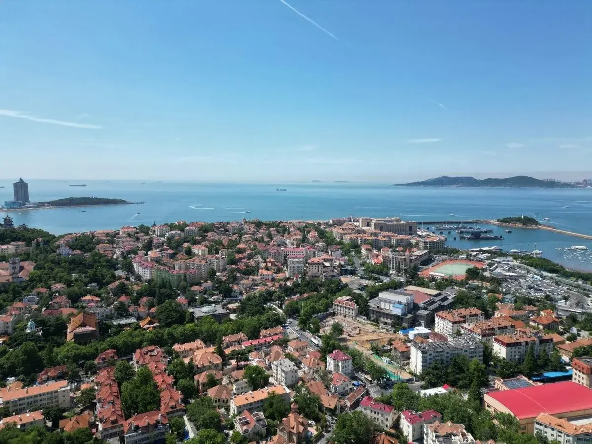 Aerial view of Qingdao's red-tile-roofed old town spread along a curving harbor, boats moored offshore and green hills behind