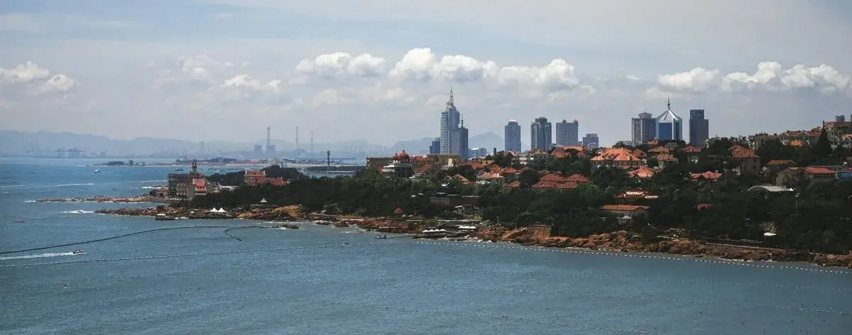 A rocky coastline in Qingdao with a distant city skyline visible across calm water under a hazy sky