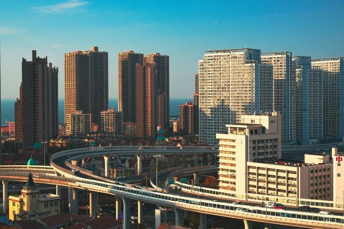 A curving elevated highway interchange in Qingdao with modern high-rise apartment towers behind it and the sea visible in the distance