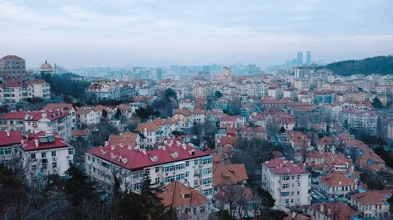 Dense red-tile-roofed housing at dusk in Qingdao's old town, with modern skyscrapers rising hazily on the horizon behind