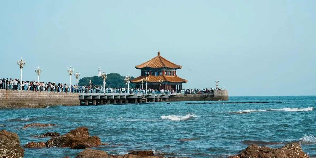 Zhan Qiao pier in Qingdao, its traditional Chinese pavilion Huilan Ge at the end, waves breaking over rocks in the foreground