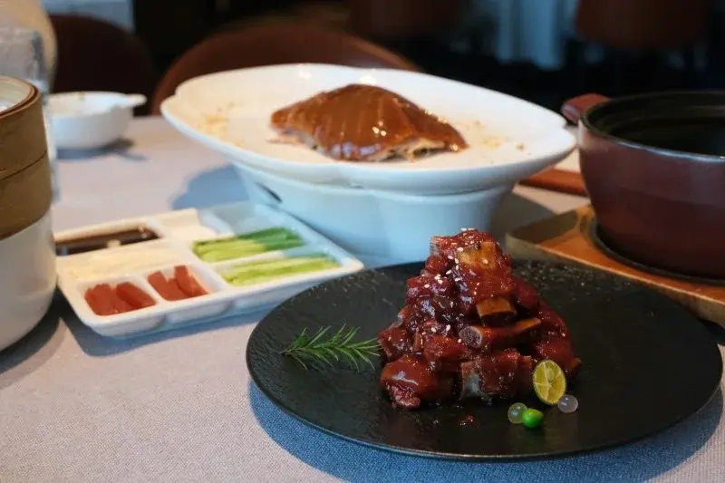 An elegantly plated red-braised pork dish and a sliced roast duck, arranged with small garnish portions on oversized plates