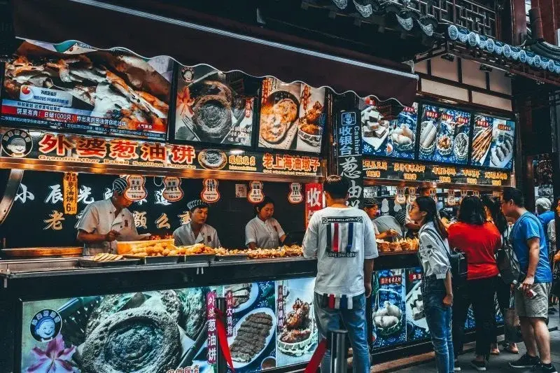 A street food stall in Shanghai with cooks preparing scallion pancakes behind a glass counter, a queue of customers waiting