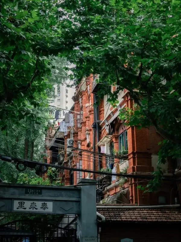 A red-brick shikumen lane house behind trees, laundry hanging from the balconies, with a modern high-rise visible in the background