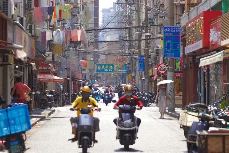 A sunny Shanghai lane with delivery riders on electric bikes, laundry overhead and a bilingual bike-lane sign